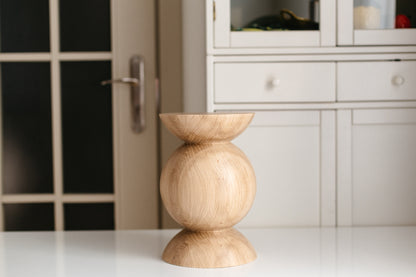 A small wooden pedestal stand with a rounded middle section and flared top and bottom bases, shown on a white tabletop with a blurred interior background featuring a cabinet and door.