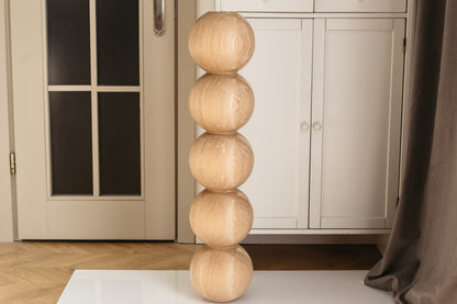 A tall, narrow decorative sculpture made of five stacked oval wooden forms with visible natural wood grain, standing on a white surface in an indoor room with a cabinet and door in the background.