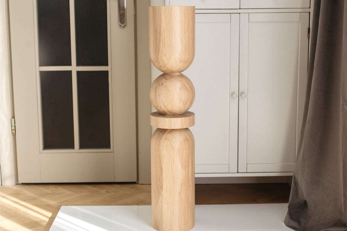 A tall, sculptural wooden vase with a stacked design of a cylinder, a sphere, and a smaller cylinder on top, made from light natural wood, standing on a white surface in a home interior.
